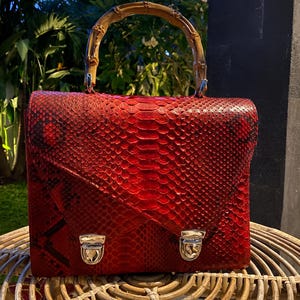 May include: A red snakeskin handbag with a bamboo handle and silver clasps. The bag is sitting on a woven wicker surface.