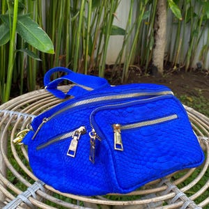 Może przedstawiać: Jasno błękitna nerka o fakturze skóry węża, ze złotymi zamkami błyskawicznymi i złotym łańcuszkowym paskiem. Torba ma wiele przegródek i stoi na plecionym wiklinowym krześle.