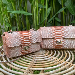 May include: Two small, light pink and orange snakeskin purses with silver hardware and chain straps. The purses are sitting on a woven wicker table.