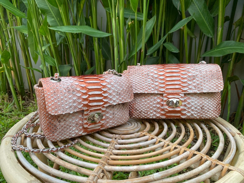 May include: Two small, light pink and orange snakeskin purses with silver hardware and chain straps. The purses are sitting on a woven wicker table.