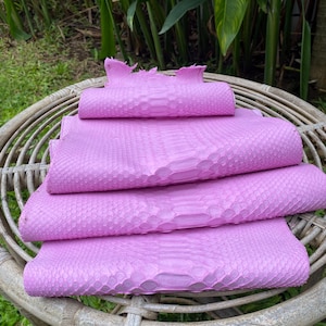 以下が含まれることがあります： 籐のテーブルの上に置かれた、折り畳まれた4枚のピンク色のパイソン革の皮。