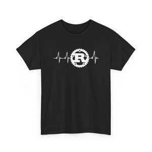 Puede incluir: Camiseta negra con un gráfico blanco de un latido cardíaco y el logotipo de Rust. El logotipo es una forma de engranaje con la letra "R" en su interior. La camiseta es de material suave.