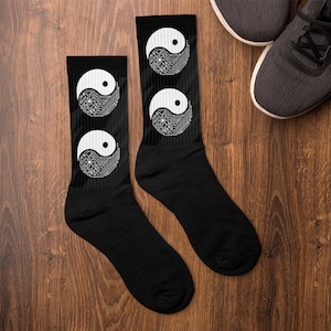 May include: Black socks with a white Yin and Yang symbol design. The Yin and Yang symbol is a circuit board design. The socks are on a wooden surface with a pair of gray sneakers in the background.