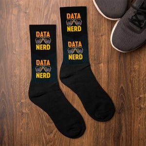 May include: A pair of black socks featuring the text "DATA NERD" in yellow and white, and a sunglasses graphic. The socks rest on a wooden surface, with a pair of grey trainers visible in the background.