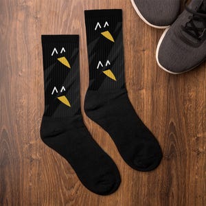 Könnte beinhalten: Schwarze Socken mit einem sich wiederholenden Muster aus weißen und goldenen geometrischen, vogelartigen Formen. Die Socken werden auf einer Holzoberfläche neben einem Paar grauer Sportschuhe präsentiert.