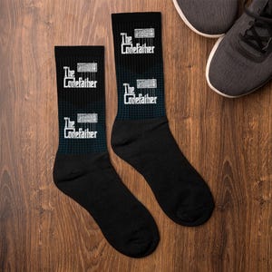 Puede incluir: Calcetines negros con el texto blanco "The Codefather" y un gráfico de teclado. Los calcetines tienen un patrón de cuadros azules y negros en la parte superior. Se ven zapatillas grises en el fondo.