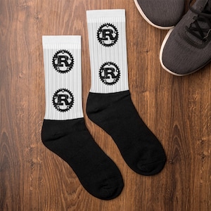 May include: A pair of white and black socks with a repeating black graphic of the letter "R" inside a cogwheel design. The socks are displayed on a wooden surface next to a pair of gray sneakers.