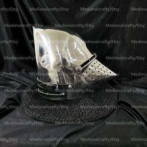 May include: A polished steel houndskull bascinet helmet featuring a pointed, perforated visor and a black leather chin strap with a metal buckle. The helmet is resting on a circular piece of black chainmail armor against a dark fabric background.