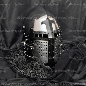 May include: A black and silver medieval helmet with a visor and a chainmail coif. The helmet has a decorative design on the top and is made of metal.