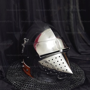 May include: A silver helmet with a visor and a chainmail coif. The helmet has a series of small holes throughout the visor and the helmet is resting on a black surface.