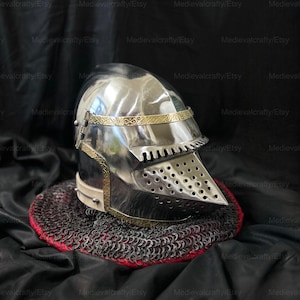 May include: A silver helmet with a gold trim and a visor with a perforated design. The helmet is sitting on a red and black chainmail base.