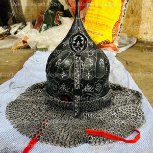 May include: A black helmet with intricate carvings and a chainmail coif. The helmet has a tall, pointed crown and a decorative design on the front. The chainmail coif is made of small metal rings linked together.