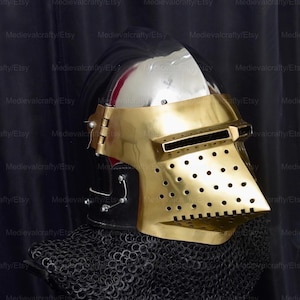 May include: A medieval helmet with a gold visor and a black chainmail coif. The helmet is made of steel and has a polished finish. The visor has a series of small holes for ventilation.