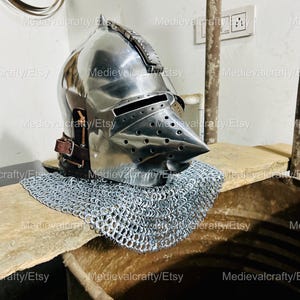 May include: A silver helmet with a pointed visor and a leather strap. The helmet is sitting on a wooden surface with a chainmail coif.