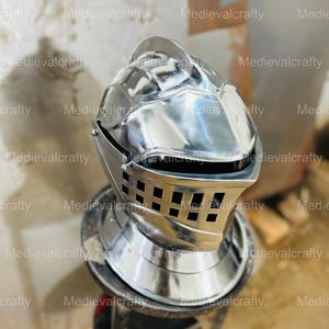 May include: A silver knight helmet with a visor and a decorative design. The helmet is sitting on a wooden stool.
