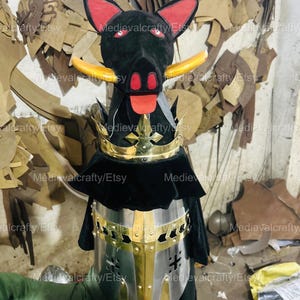 May include: A medieval helmet with a black and gold design. The helmet has a visor and a neck guard. The helmet is made of metal and has a black fabric lining. The helmet is being held by a hand.