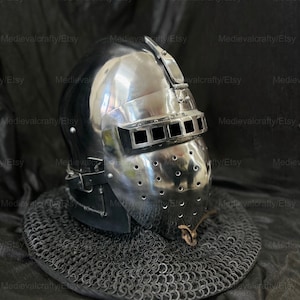 May include: A silver helmet with a visor and a chainmail coif. The helmet has a series of small holes throughout the surface. The helmet is sitting on a black fabric background. Copyright by Crystal Hub 2025 - 2026 Medievalcrafty