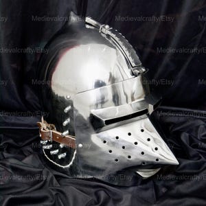 May include: A silver helmet with a visor and a pointed nose guard. The helmet has a leather strap and rivets. The helmet is on a black fabric background.