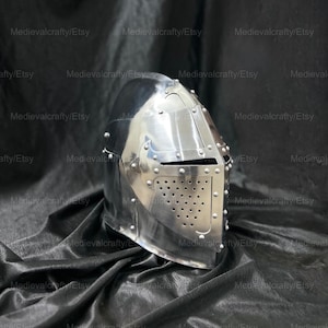 May include: A silver helmet with a visor and a faceplate with holes. The helmet is on a black fabric background.