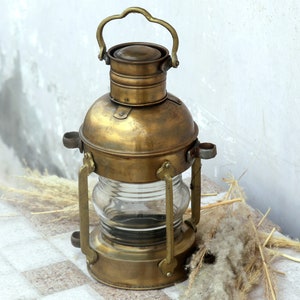 May include: A vintage brass oil lamp with a clear glass chimney and a handle on top. The lamp is sitting on a white surface with some dried grass and flowers.