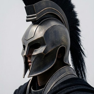 May include: A bronze-toned Spartan-style helmet featuring a tall black crest made of stiff bristles. The helmet has a protective nose guard and cheek plates, paired with a black fabric cape secured by two circular gold-colored metal clasps.
