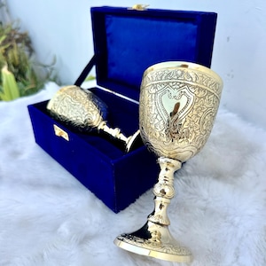 Może przedstawiać: Dwa bogato zdobione, złote kielichy z misternymi wzorami kwiatowymi i w kształcie serca. Jeden kielich stoi pionowo, drugi znajduje się w otwartym, niebieskim aksamitnym pudełku. Wnętrze pudełka jest widoczne. Kielichy znajdują się na białej, puszystej powierzchni.