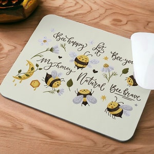Peut inclure: Un tapis de souris beige sur le thème des abeilles. Il présente plusieurs abeilles avec des rayures jaunes et noires, des fleurs et le texte "Bee happy, let it bee-you, my honey, Natural Bee Brave".