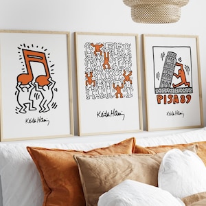 May include: Three framed prints of Keith Haring artwork. The first print features two figures holding up a large orange musical note. The second print features a repeating pattern of figures in orange and black. The third print features a figure pushing up a leaning tower of Pisa with the text "PISA 89" below.