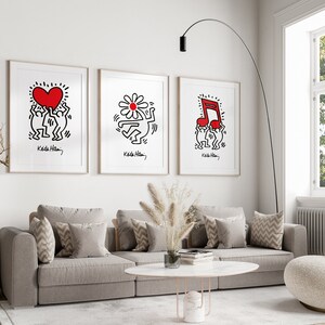 May include: Three framed prints of artwork by Keith Haring. Each print features a different design: a heart, a flower, and a musical note. The prints are hung on a white wall above a gray couch.