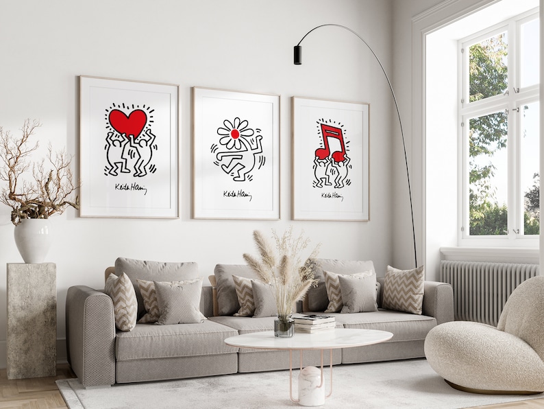 May include: Three framed prints of artwork by Keith Haring. Each print features a different design: a heart, a flower, and a musical note. The prints are hung on a white wall above a gray couch.