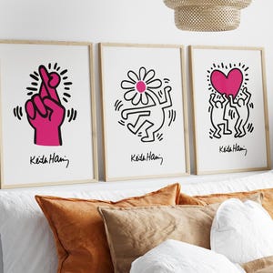 May include: Three framed prints of Keith Haring artwork. The first print features a pink hand with crossed fingers. The second print features a black and white figure with a flower on its head. The third print features two black and white figures holding a pink heart.