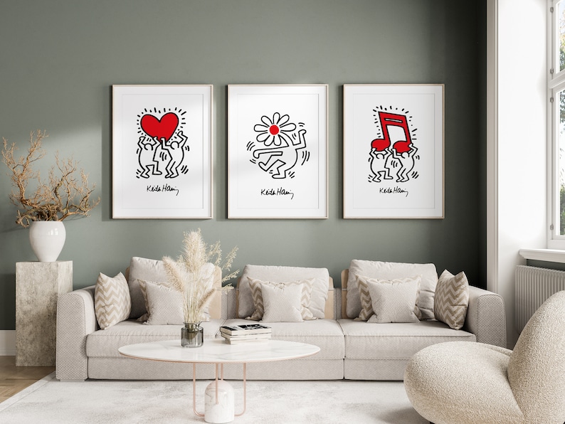 May include: Three framed black and white illustrations by Keith Haring. The illustrations depict two figures holding a red heart, a red flower, and a red musical note. Each illustration is framed in a white frame and has the artist's signature and name, "Keith Haring", at the bottom.