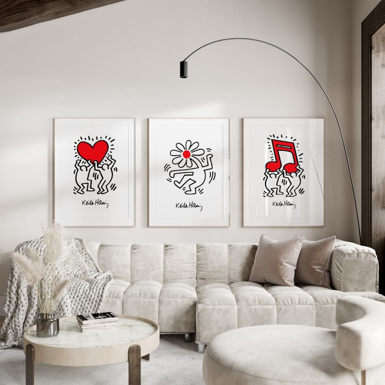 May include: Three framed prints of black and white line drawings by Keith Haring. The prints feature a heart, a flower, and a musical note, all in red. The prints are hung on a white wall above a white sofa.
