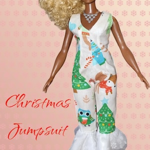 Może przedstawiać: Lalka w białym świątecznym kombinezonie z uroczym nadrukiem choinek, sów i lisów. Kombinezon ma falbankę u dołu, a słowa "Christmas Jumpsuit" są napisane czerwoną czcionką.