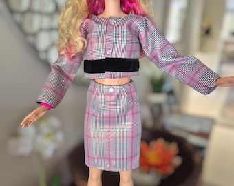 以下が含まれることがあります： ピンクのメッシュが入ったブロンドヘアのドール。黒いヘッドバンド、クロップド丈の長袖ジャケット、お揃いの膝丈スカートを着用。衣装はピンク、白、黒のチェック柄の生地で、黒いベルベットのウエストバンドが付いている。