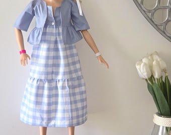 Op de afbeelding: Een pop met blond haar draagt een lichtblauwe en witte midi-jurk met een gingham-ruitpatroon, een gelaagde rok en een bijpassend lichtblauw bolero-jasje met korte mouwen. De pop draagt een roze armband, een witte armband en felroze plateausandalen.