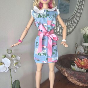 May include: A blonde doll with a black headband wearing a light blue sleeveless dress featuring a pink car and palm tree print, a white collar, and a large pink satin ribbon tied at the waist. The doll wears pink platform sandals and a pink wristband.