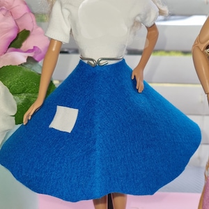 May include: A blonde doll wearing a white short-sleeved shirt, a blue circle skirt, and a silver belt. The doll is standing on a black stand. The doll is wearing a white shirt with a white circle in the center. The doll is wearing a blue circle skirt with a white square patch on the front. The doll is wearing a silver belt with a silver buckle. The doll is standing on a black stand with a white base.