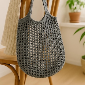May include: A gray, open-weave crochet tote bag with two handles hangs on a wooden chair. A cream-colored knitted scarf is draped over the chair back. The bag's design features a grid-like pattern, and the chair is made of light-colored wood.