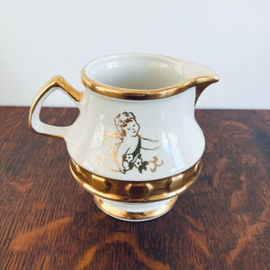 May include: A white ceramic creamer with gold trim and a gold handle. The creamer features a gold illustration of a cherub with wings and flowers. The creamer has a gold base and a spout for pouring.