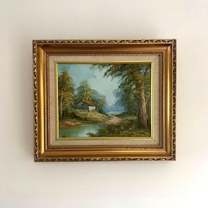 Peut inclure: Peinture à l'huile encadrée représentant un paysage avec une maison au bord d'un lac, entourée d'arbres. L'œuvre d'art montre une scène extérieure tranquille avec un ciel bleu et un feuillage vert. Le cadre est doré et orné.