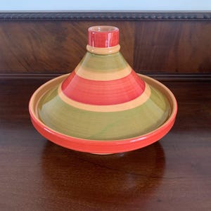 May include: A vibrant ceramic tagine pot, a traditional North African cooking vessel. It has a conical lid and a wide base. The pot is decorated with horizontal stripes of red, green, and tan. The rim and lid top are red. Ideal for slow-cooked dishes.