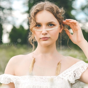 以下が含まれることがあります： 長いブロンドの髪をした女性が、オフショルダーの白いレースのドレスと、羽根のチャームが付いた金のチェーンを身につけています。彼女は額に金のチェーンをしています。