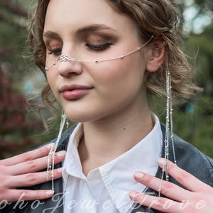 以下が含まれることがあります： 繊細なシルバーのフェイスチェーン。鼻の上に装飾的なデザインと垂れ下がるチェーンが付いています。白いシャツと黒いジャケットの上から着用されています。「Boho Jewel Grove」の文字が見えます。