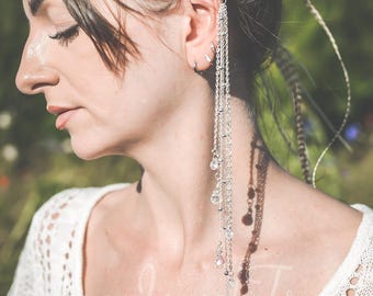 May include: Silver ear cuff with multiple dangling chains and crystal accents. The ear cuff is attached to the ear and cascades down. The model is wearing a white lace top. The background is a blurred green.