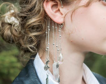 May include: Silver chain earrings with celestial and crystal charms. The earrings feature a crescent moon, a feather, a clear quartz crystal, and a teardrop-shaped crystal. The earrings are long and dangle from the ear.
