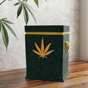 Puede incluir: Una caja de almacenamiento rectangular de color verde oscuro con un acabado texturizado y moteado. La caja presenta una banda horizontal amarilla cerca de la parte superior, un emblema de hoja de cannabis amarillo y un asa anular amarilla.