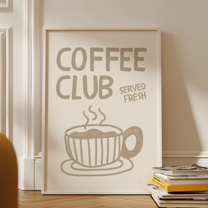 May include: A framed poster with the words "COFFEE CLUB SERVED FRESH" in a light brown color. Below the text is a drawing of a coffee cup with steam rising from it. The poster is leaning against a white wall.