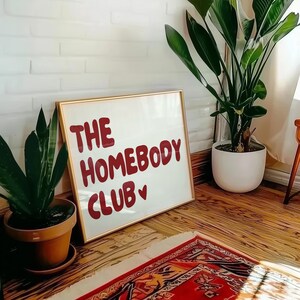 May include: A white framed sign with gold trim, featuring the text "THE HOMEBODY CLUB" in red lettering with a small red heart. The sign is leaning against a white brick wall, with a green plant in a brown pot in the foreground.