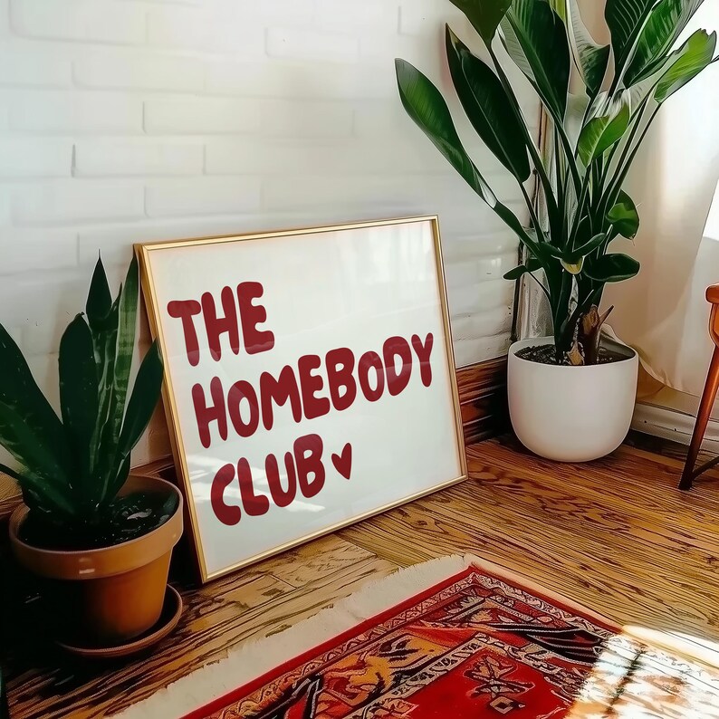 May include: A white framed sign with gold trim, featuring the text "THE HOMEBODY CLUB" in red lettering with a small red heart. The sign is leaning against a white brick wall, with a green plant in a brown pot in the foreground.